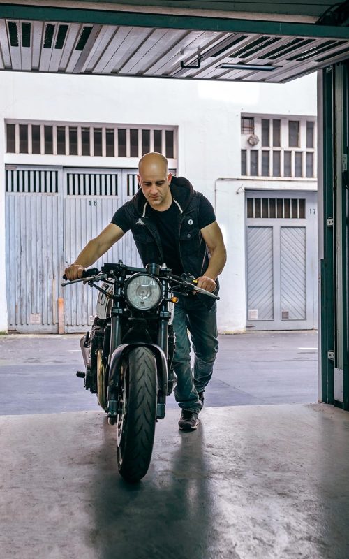 Man pushing a black motorcycle through an open garage doorway.