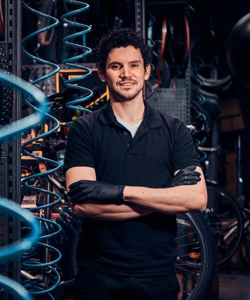 Smiling mechanic in a black shirt and gloves with folded arms in a workshop.